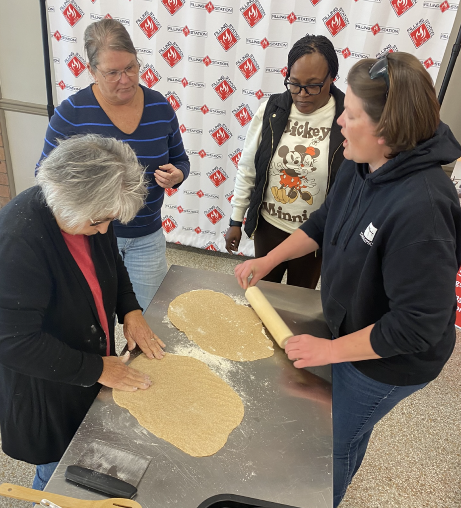 Women stand around a table rolling out pizza dough.